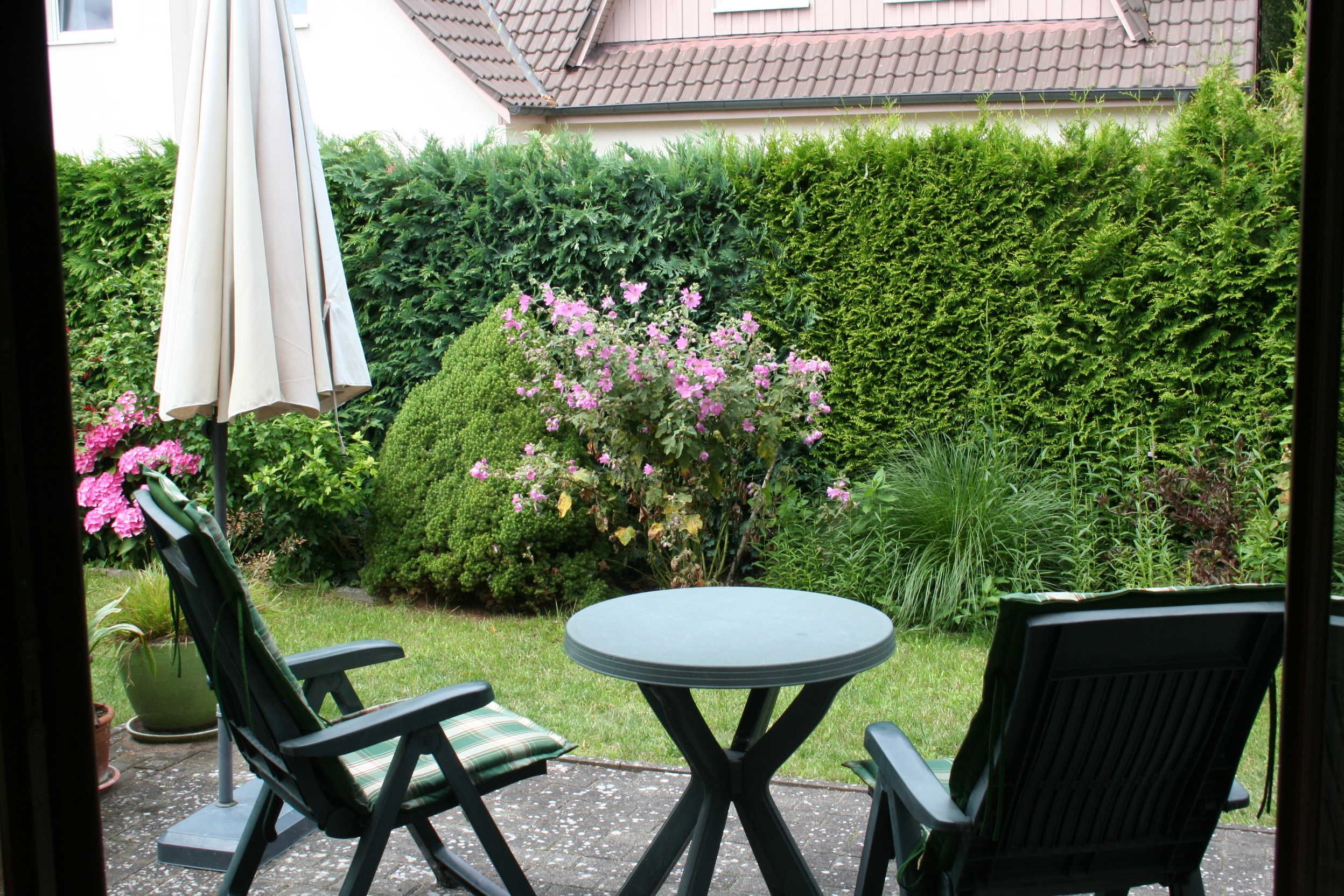 Une terrasse avec deux chaises et une table, entourée d’une haie et de plantes en fleurs.