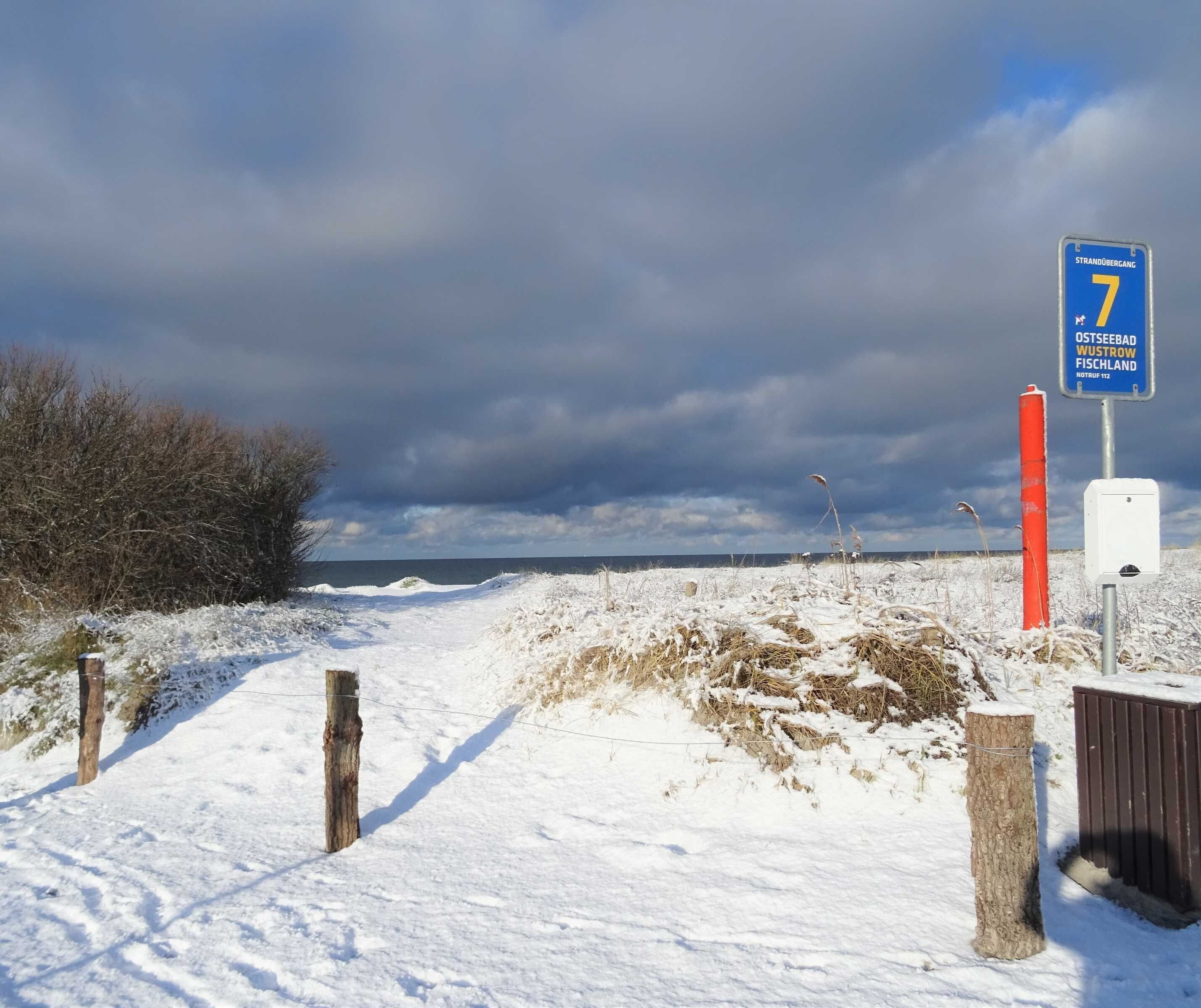 Un paysage enneigé avec un sentier menant à la mer, un panneau indiquant « Strandbergang 7 », et un poteau rouge.