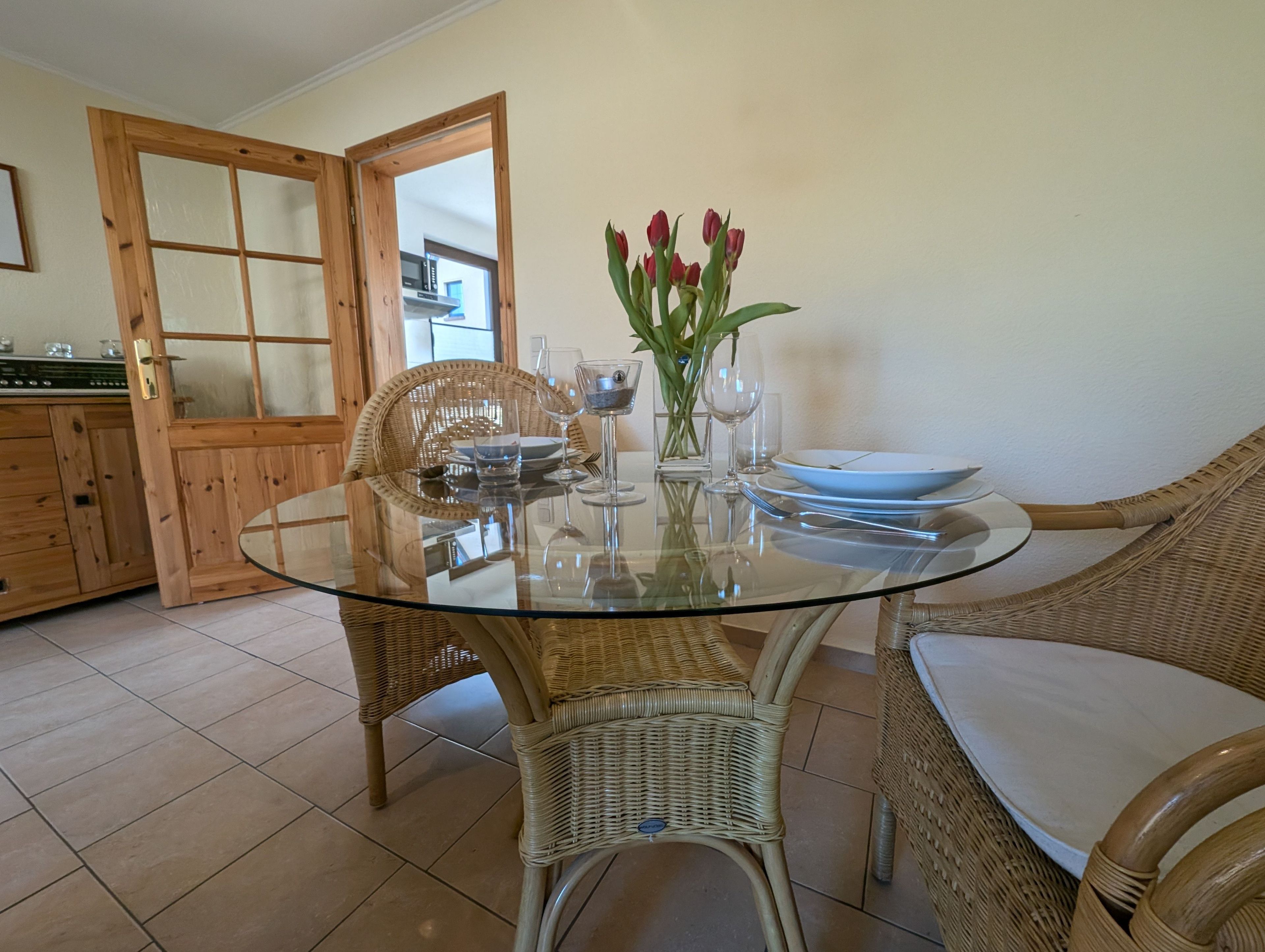 Un coin repas avec une table en verre, des chaises en osier, un vase rempli de fleurs rouges et, au fond, une porte en bois.