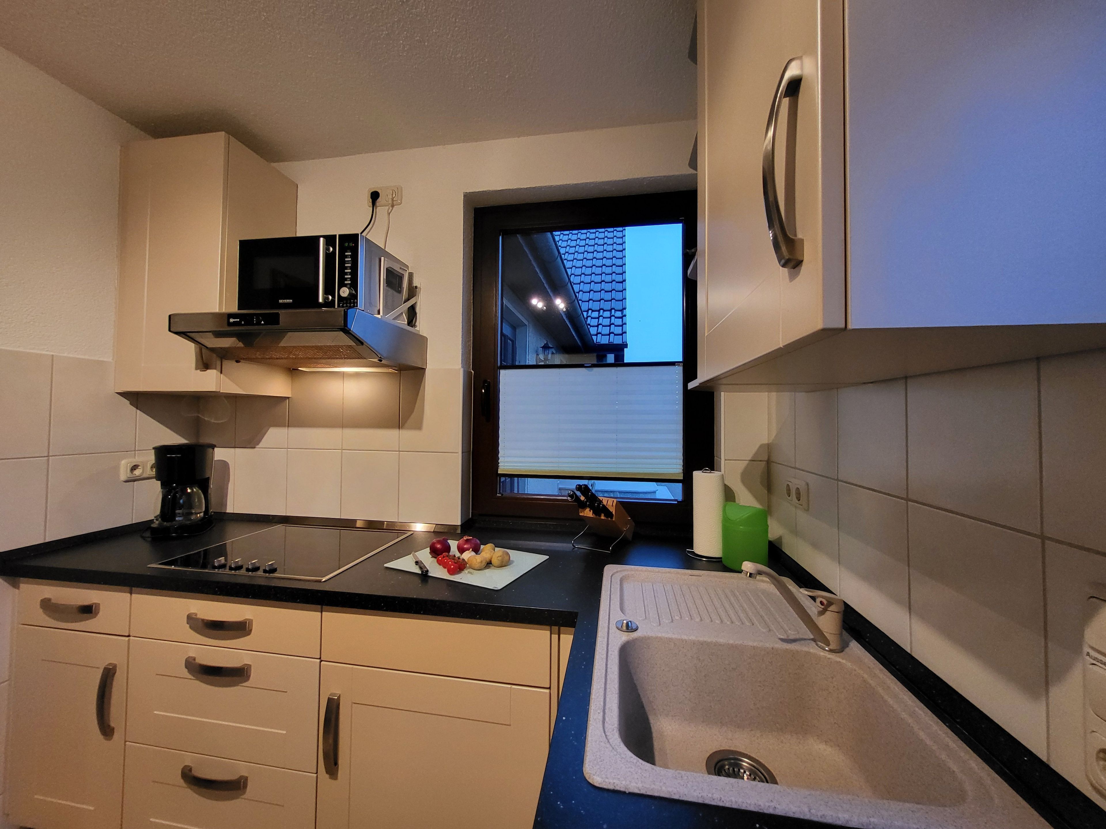Ett kök med mikrovågsugn, kaffebryggare, diskho och ett fönster med utsikt över en byggnad.