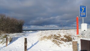 Un paysage enneigé avec un sentier menant à la mer, un panneau indiquant « Strandbergang 7 », et un poteau rouge.