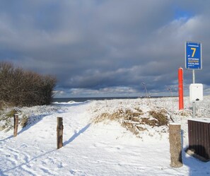 Lumipeitteinen maisema, jossa polku johtaa merelle, kyltti, jossa lukee ”Strandbergang 7”, ja punainen tolppa.