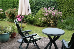 Une terrasse extérieure avec une table ronde et deux chaises, entourée de plantes en pot et d’arbustes en fleurs.