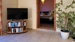 Un salon avec un téléviseur posé sur un meuble, une bibliothèque, une plante en pot et une porte donnant sur une autre pièce.