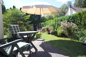 Een terras buiten, met een tafel en stoelen, omringd door weelderig groen en bloeiende planten.
