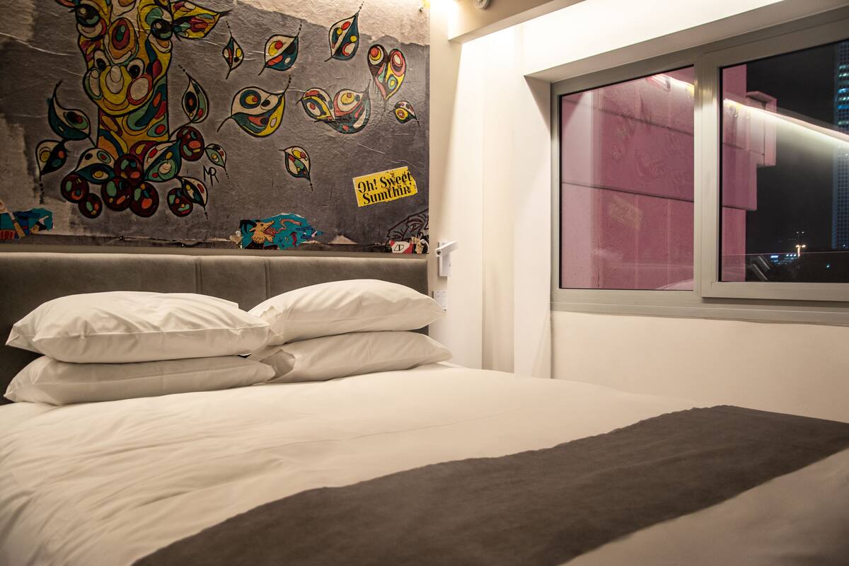 Ein Bett mit weißen Leinen und einer grauen Tagesdecke, eine Wand mit farbenfroher abstrakter Kunst und ein Fenster mit rosafarben getöntem Glas.