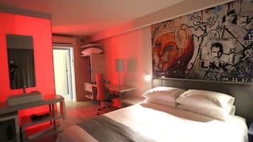 A hotel room with a bed, desk, chair, and a wall with graffiti art.