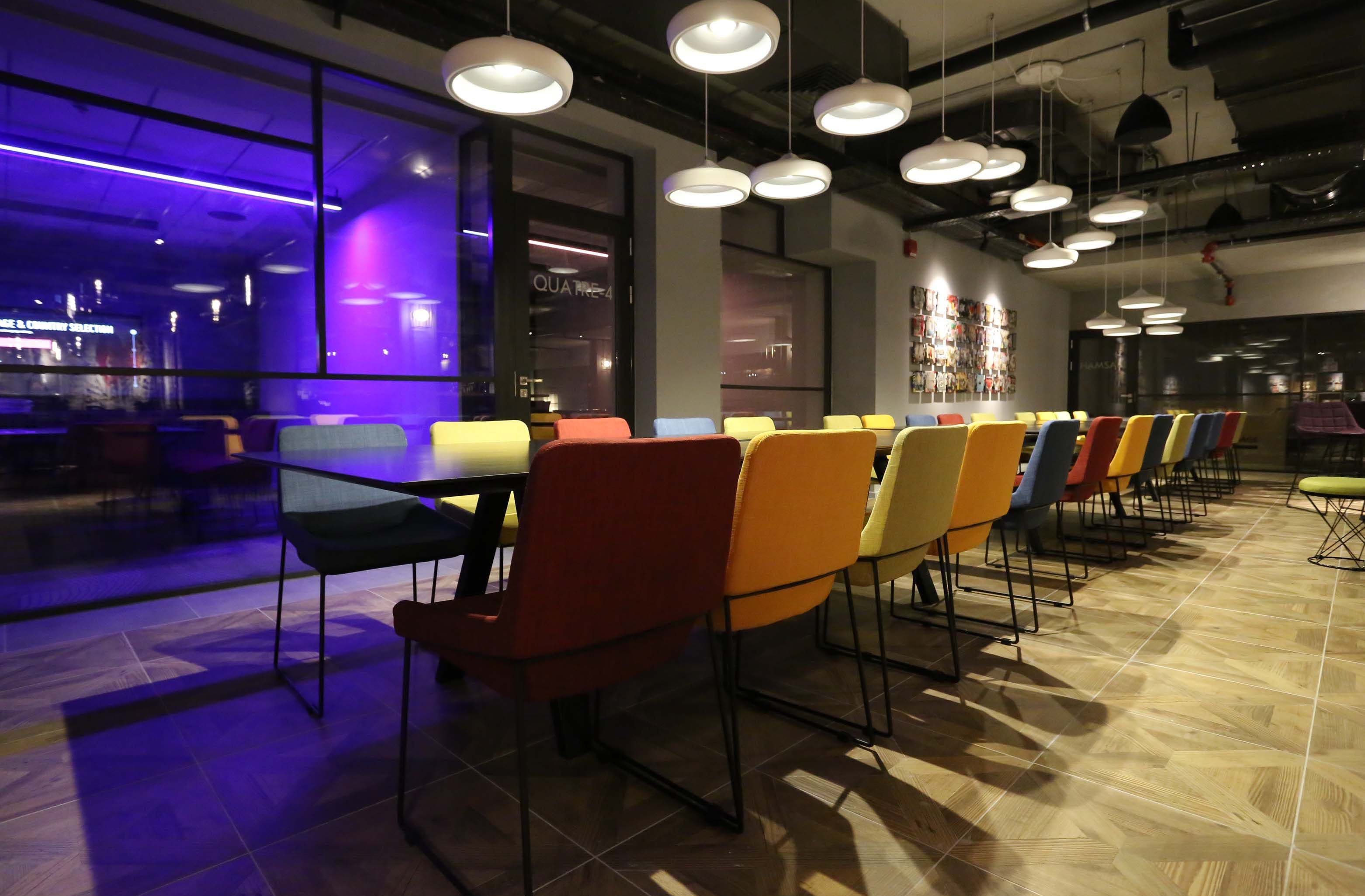 Un comedor moderno con una mesa larga y sillas de colores, iluminación colgante y una cocina visible al fondo.