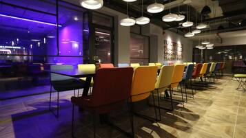 Un espace de salle à manger moderne avec une longue table et des chaises colorées, un éclairage suspendu et une cuisine ouverte visible au fond.
