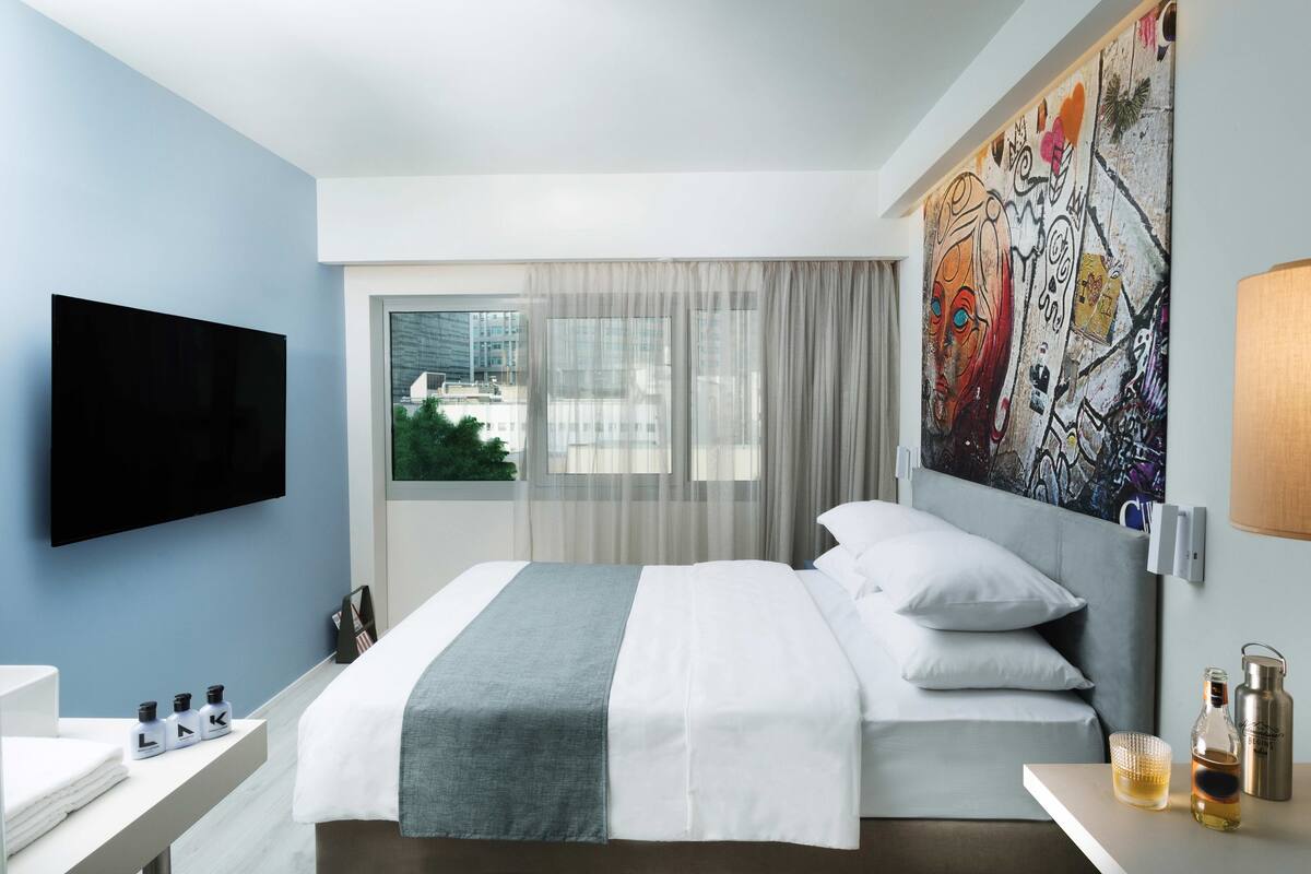Ein modernes Hotelzimmer mit einem großen Bett, einem Flachbildfernseher, einem Fenster mit transparenten Vorhängen und einem farbenfrohen abstrakten Gemälde an der Wand.