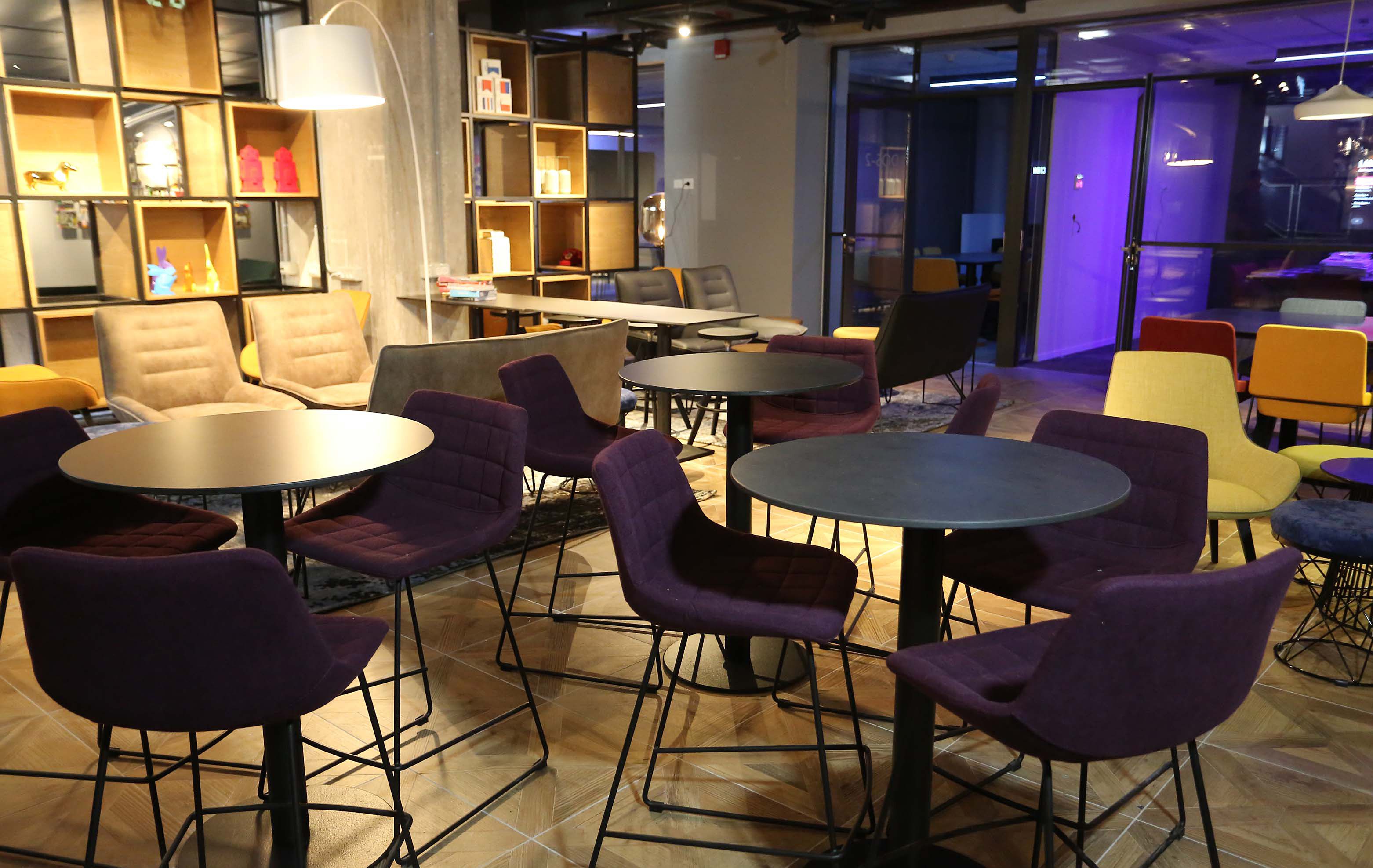 A modern lounge area with round tables and purple chairs, wooden shelves with various items, and a view through a glass door.