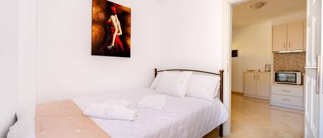 A bedroom with a bed, white walls, a painting, and a view into the kitchen.