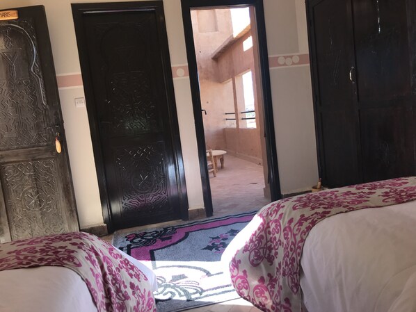 A hotel room with two beds, a wooden door, and a view of an outdoor area with a chair and a table. - Kasbah Rayane Ait Ben Haddou (Aït Benhaddou)