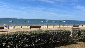 A beach with benches, a hedge, and boats on the water.