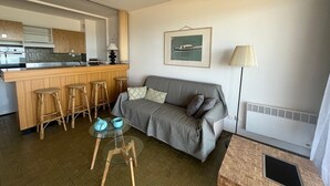 A modern living space with a kitchen, a grey sofa, a small round table with stools, and a lamp.