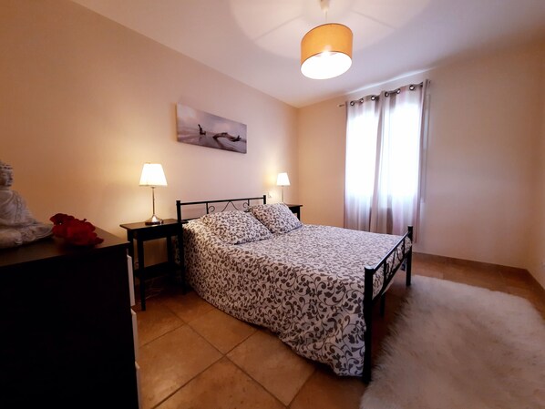 A bedroom with a bed, two bedside tables with lamps, a wall-mounted painting, and a window with curtains.