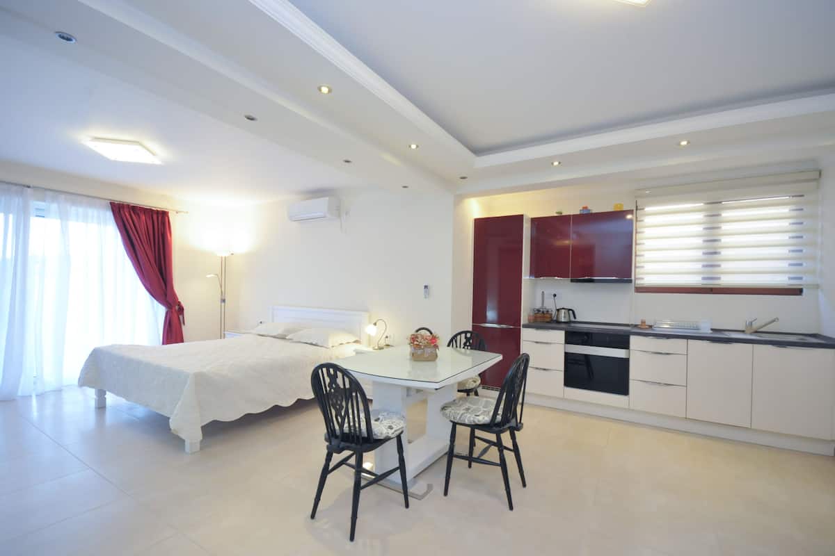 A modern hotel room with a bed, a dining table with chairs, a kitchen area, and a large window with blinds.