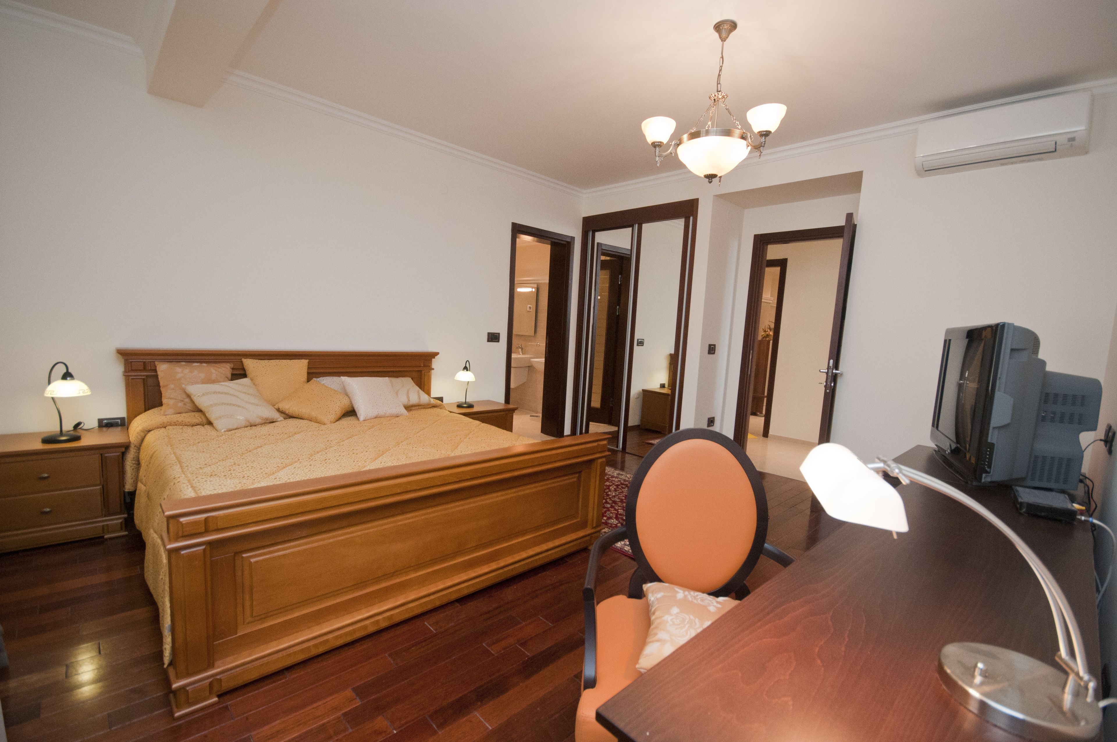 Ein Schlafzimmer mit einem hölzernen Bett, Nachttischlampen, einem Schreibtisch mit Stuhl, einem Fernseher und einem Spiegel.