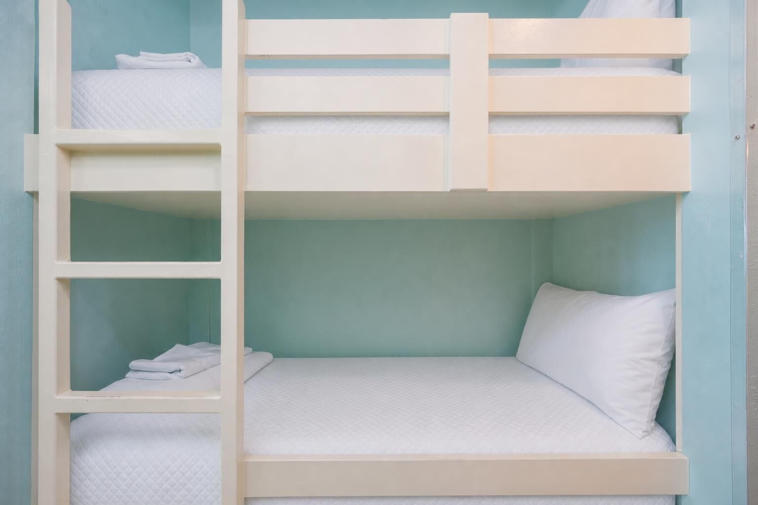 A bunk bed with a white mattress and pillows, a white ladder, and a teal wall.