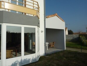 Ein modernes, zweistöckiges Haus mit Balkon, großen Schiebeglastüren und einer kleinen Terrasse mit Gartenmöbeln.