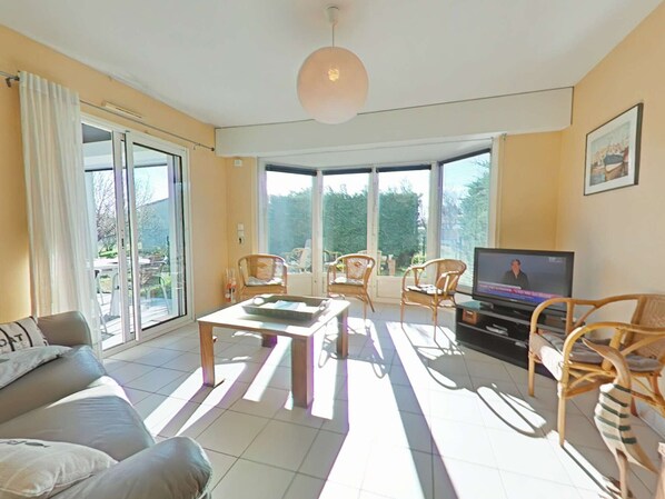 A spacious living room with a sofa, a coffee table, and a TV. There are wicker chairs and a large sliding glass door leading to an outdoor area.
