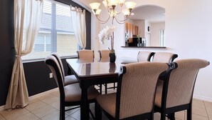 A dining area with a dark wood table and chairs, a chandelier, and a view of the kitchen through a window with curtains.