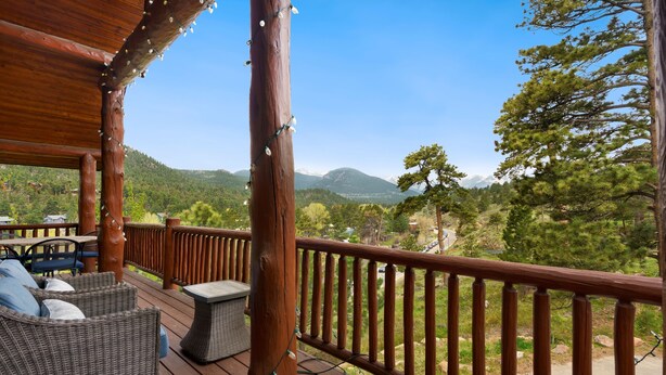 Una terraza de madera con mobiliario de mimbre y barandilla, con vista a un paisaje montañoso.