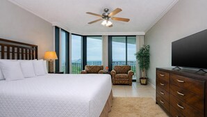 A bedroom with a bed, a television, a ceiling fan, a chair, a plant, and a view of the outdoors.