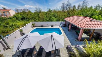 A rooftop pool area with lounge chairs and umbrellas, surrounded by greenery and a building with a red roof.