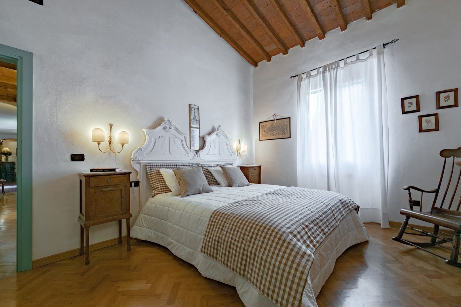 A bedroom with a large bed, wooden headboard, two bedside tables, a nightstand, a chair, and a window with curtains.
