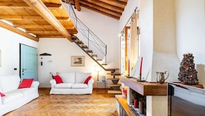 Un vaste salon doté d’un parquet en bois disposé en point de Hongrie, de canapés blancs agrémentés de coussins rouges, d’une cheminée, d’un téléviseur et d’un escalier menant à un niveau supérieur.