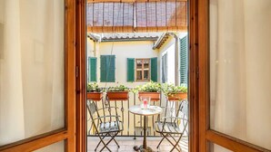 On aperçoit, à travers une porte à cadre en bois, une cour intérieure avec une table et deux chaises, entourée de plantes en pot et d’un bâtiment aux volets verts.