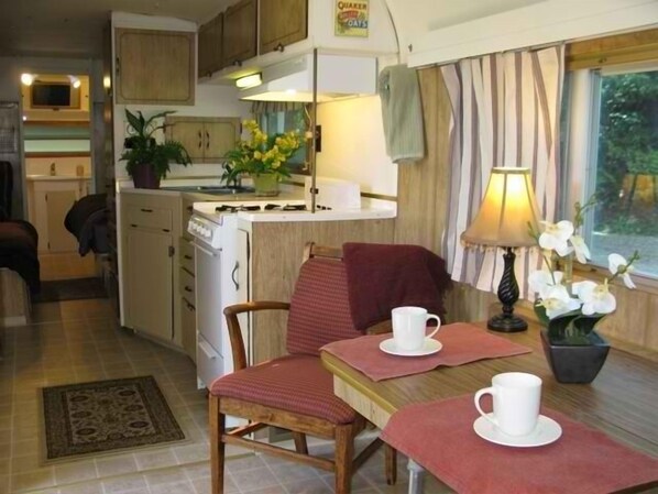 A compact kitchen with a stove, sink, and cabinets, a dining area with a table and chairs, and a window with curtains.