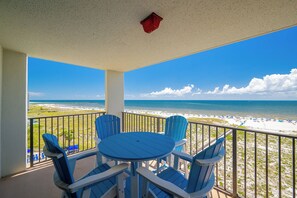 A balcony with a table and chairs, overlooking a beach with a clear view of the ocean and a sandy shore.