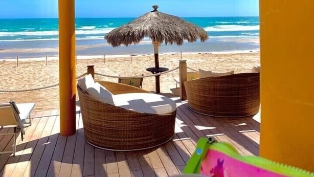 A beachside patio with wicker furniture, a thatched umbrella, and a view of the ocean.