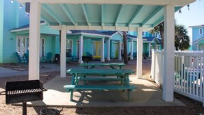 A covered outdoor area with picnic tables and a grill, surrounded by colorful houses with a white fence. - Pet Friendly, Located in Town, Close to beach, Shared Pool, Golf Cart Access (Port Aransas)