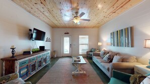 A cozy living room with a wooden ceiling, a ceiling fan, a television mounted on the wall, a sofa, a coffee table, and a cabinet with drawers. - Pet Friendly, Located in Town, Close to beach, Shared Pool, Golf Cart Access (Port Aransas)