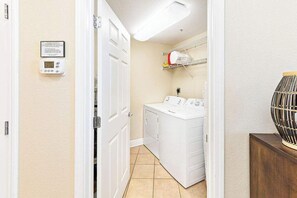 A laundry room with a washing machine and dryer, a wall-mounted thermostat, and a shelf with laundry supplies. - Crystal Tower 1509 - Luxurious 15th Floor Corner Condo with Breathtaking Views (Gulf Shores)