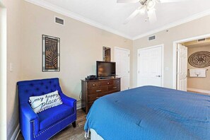Une chambre avec un grand lit, un fauteuil bleu, une commode avec un téléviseur et un élément décoratif mural.