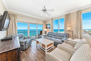 A spacious living room with a large sofa, a coffee table, and a TV. There are large windows offering a view of the ocean. - Crystal Tower 1509 - Luxurious 15th Floor Corner Condo with Breathtaking Views (Gulf Shores)