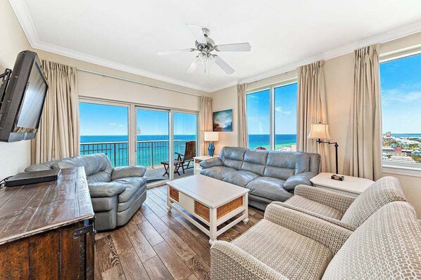 A spacious living room with a large sofa, a coffee table, and a TV. There are large windows offering a view of the ocean.