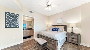 Een slaapkamer met een houten bed, nachtkastje, een bankje, een plafondventilator en uitzicht op de keuken.