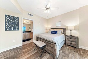 A bedroom with a wooden bed, a nightstand, a bench, a ceiling fan, and a view into the kitchen. - Crystal Tower 1509 - Luxurious 15th Floor Corner Condo with Breathtaking Views (Gulf Shores)