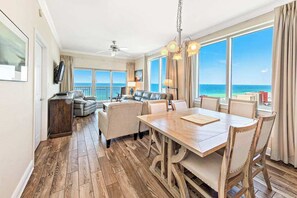 A spacious living area with a wooden floor, a dining table, and a sofa set. Large windows offer a view of the ocean.
