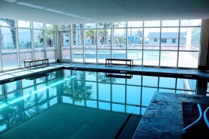 Une grande piscine intérieure aux parois en verre transparent, avec des bancs le long des côtés et une vue sur les palmiers et les bâtiments à l’extérieur.
