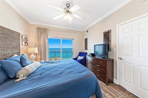 A bedroom with a bed, a television, a dresser, a ceiling fan, and a view of the ocean through a balcony door.