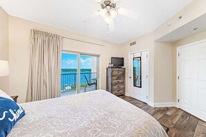 A bedroom with a large bed, a ceiling fan, a TV, and a view of the ocean through a sliding door.