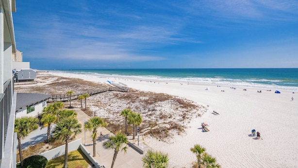 Blick auf einen Strand vom Gebäude aus, mit einer Promenade, Palmen und blauem Himmel.