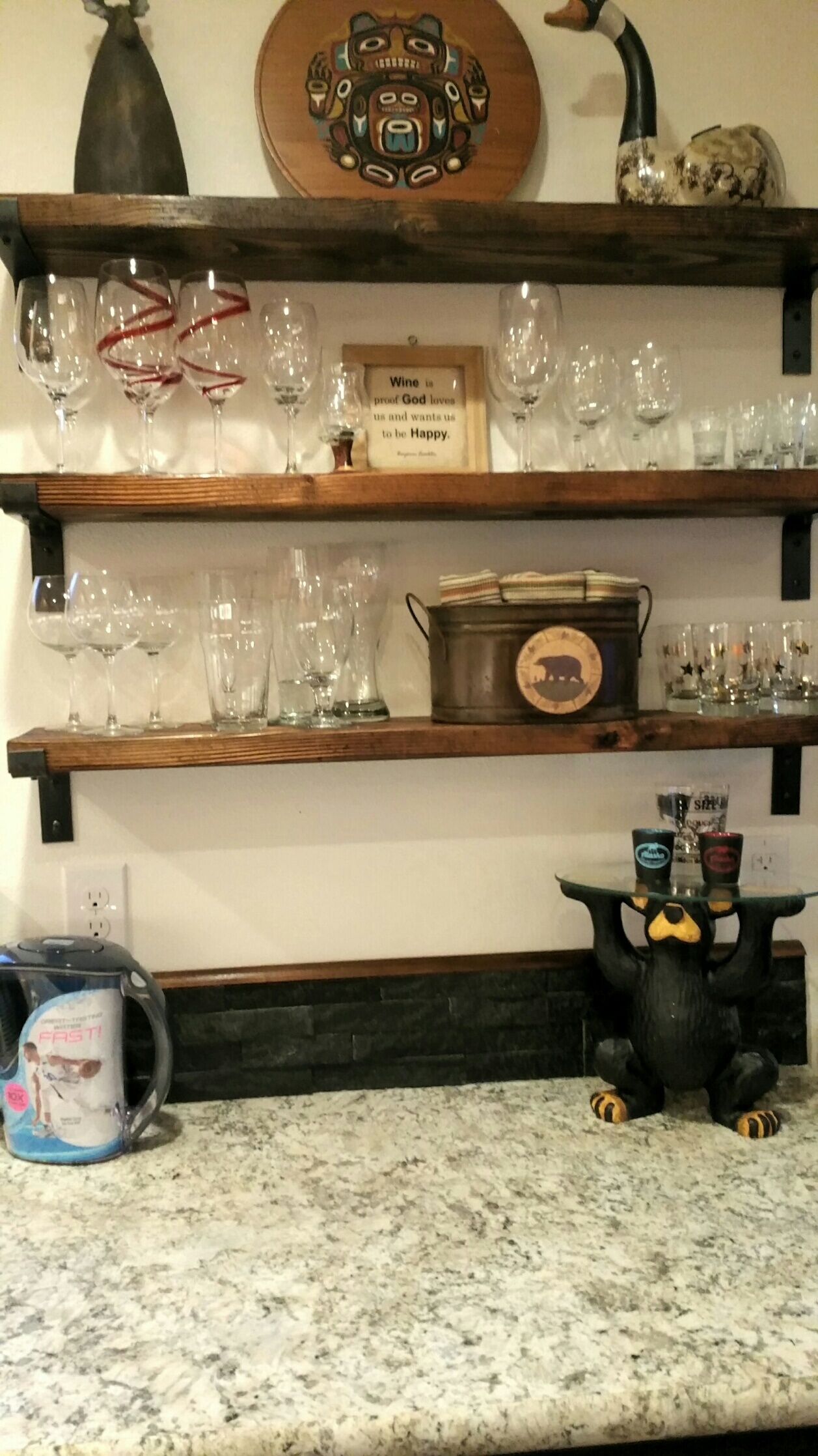 A kitchen counter with a coffee maker, a decorative bear figurine, and shelves with wine glasses and a metal pot.