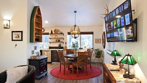 Een eethoek met een houten tafel, stoelen en een rood kleed. Daarboven hangt een kroonluchter en aan de muur is een televisie te zien, waarop verschillende zenders worden vertoond.